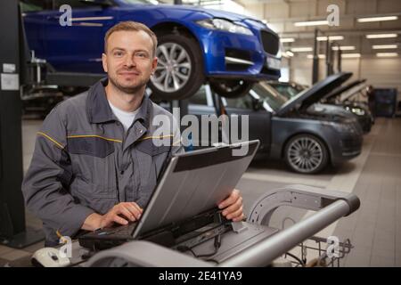Cheerful mature male car mechanic smiling to the camera, working on a computer at his garage. Happy auto technician repairing automobile at his worksh Stock Photo