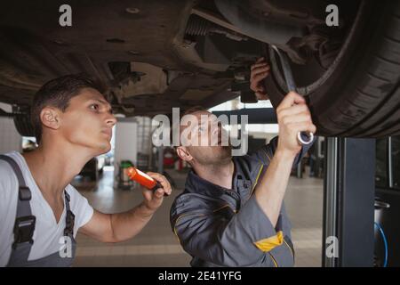 Two car mechanics working underneath a broken vehicle at the garage. Experienced auto technicians making adjustments on underside of an auto Stock Photo