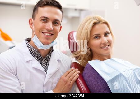 Gorgeous mature woman and her male dentist smiling happily to the camera at the dental clinic. Professional dentist and his satisfied client after den Stock Photo