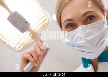 Close up of a mature female dentist smiling, wearing medical mask, adjusting dental lamp. Professional dentist preparing for teeth examination, lookin Stock Photo
