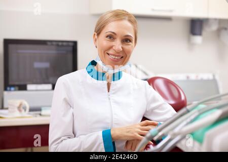 Happy mature female dentist smiling to the camera, posing at her office. Professional dentist waiting for her patient for dental examination. Dentistr Stock Photo