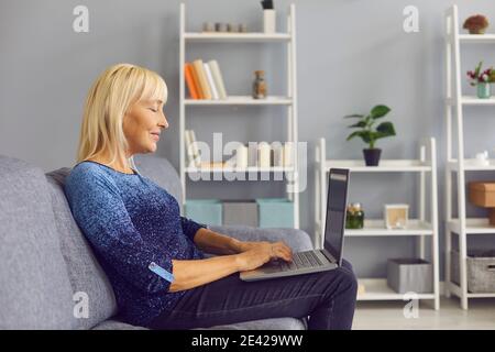 Side view of a senior woman typing an email on a laptop while sitting on her couch at home. Stock Photo