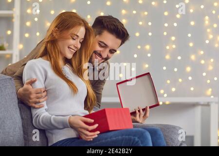 Side view of a woman with her husband opening a gift box while sitting at home on the couch. Stock Photo