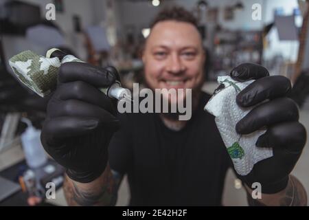 Close up of a happy bearded tattoer smiling, reaching out to the camera holding tattoo machine. Point of view shot of a tattooer holding out tattoo ma Stock Photo