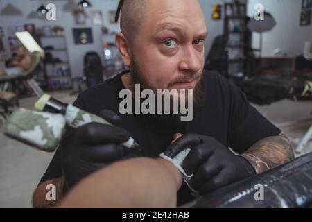 Cropped fish eye close up of a professional tattoo artist looking suspiciously to the camera, raising his brow while tattooing customer Stock Photo