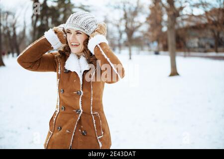 happy stylish woman with mittens in a knitted hat and sheepskin coat looking into the distance outdoors in the city park in winter. Stock Photo