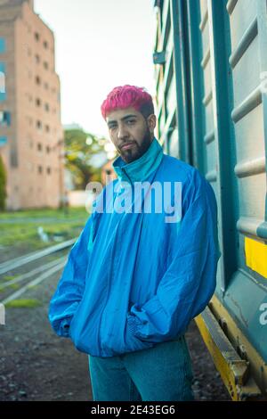 Fashionable young man with stylish pink-dyed hair wearing a retro jacket posing near a railway wagon Stock Photo