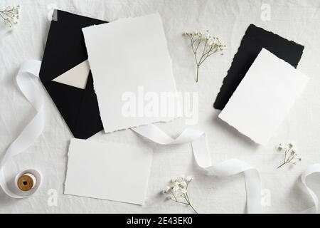 Black envelopes and white paper cards with flowers and ribbon on textile background. Flat lay, top view. Wedding flat lay composition. Stock Photo