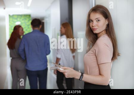 Attractive young businesswoman smiling to the camera, her colleagues walking the corridor on background Stock Photo