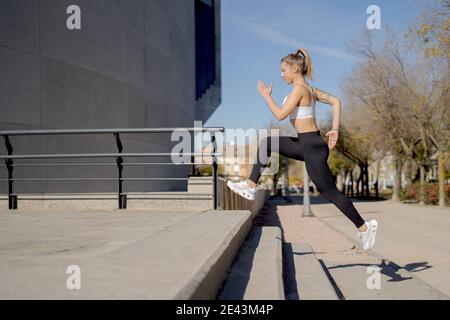 Full body side view of energetic young fit female in sportswear running fast upstairs during fitness workout on urban street Stock Photo