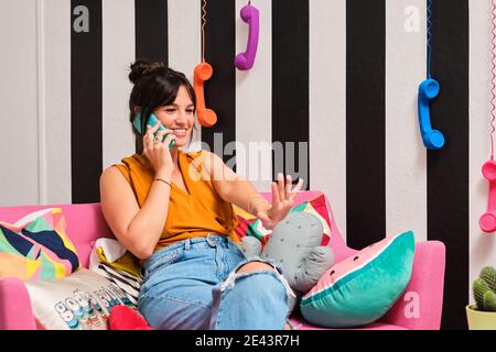 Happy young Woman with outstretched arm having phone conversation and discussing new manicure with friend while resting on sofa in stylish room with c Stock Photo