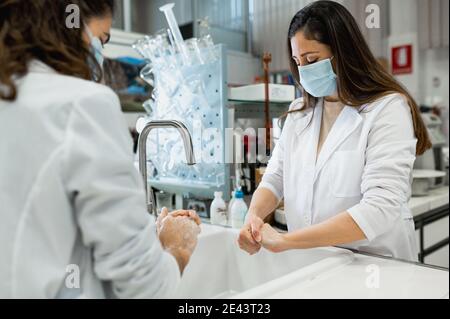 Side view of female scientists in white coats and protective masks washing hands before conducting chemical experiment in modern laboratory Stock Photo