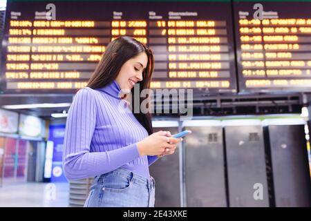 Side view of happy young ethnic female traveler in casual clothes standing in modern railway station near train schedule board and and smiling while m Stock Photo