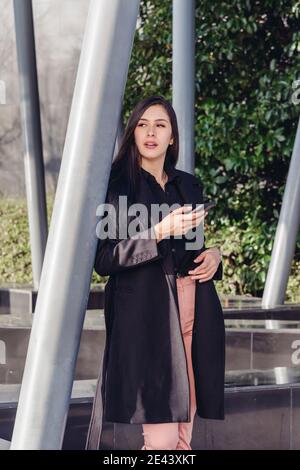 Young female in stylish outfit standing leaning on metal railing of modern building on terrace and browsing mobile phone while checking information fo Stock Photo