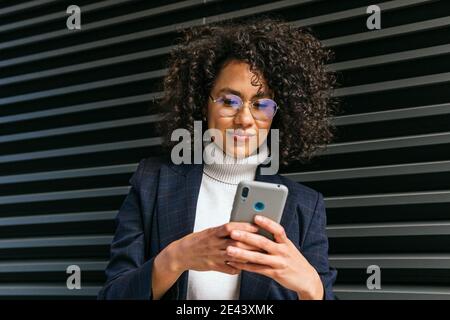 Smiling young ethnic curly haired female in stylish outfit browsing on mobile phone against gray wall Stock Photo