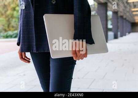 Cropped unrecognizable successful well dressed female in elegant formal wear with laptop in hands while standing near contemporary business building Stock Photo