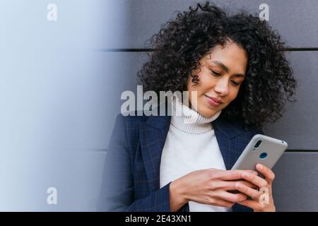 Positive young curly haired ethnic businessWoman reading information on mobile phone while standing near gray wall Stock Photo