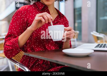 Cropped unrecognizable ethnic businessWoman sitting at table drinking coffee while working remotely on project on laptop Stock Photo