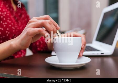 Cropped unrecognizable ethnic businessWoman sitting at table drinking coffee while working remotely on project on laptop Stock Photo