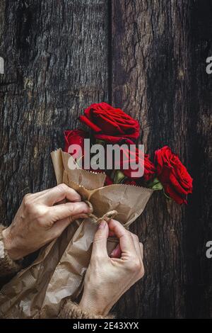 Top view of crop anonymous female in warm sweater wrapping bouquet of red roses with craft paper and rope against wooden background Stock Photo