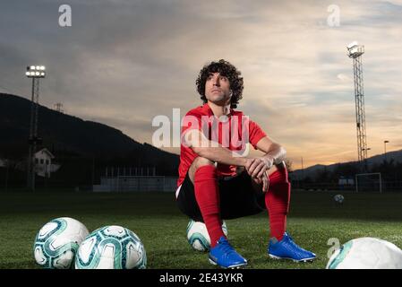 Low angle full body of serious determined sportsman in red soccer uniform sitting on field with football balls and looking away thoughtfully while res Stock Photo