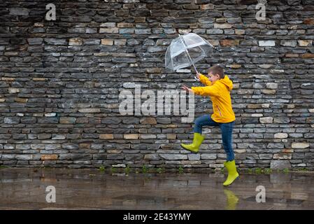 Side view of carefree male child in yellow raincoat and boots jumping under umbrella along wet street on rainy day in city Stock Photo