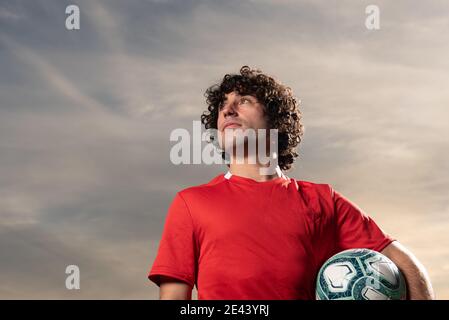 Low angle full body of serious determined sportsman in red soccer uniform sitting on field with football balls and looking away thoughtfully while res Stock Photo