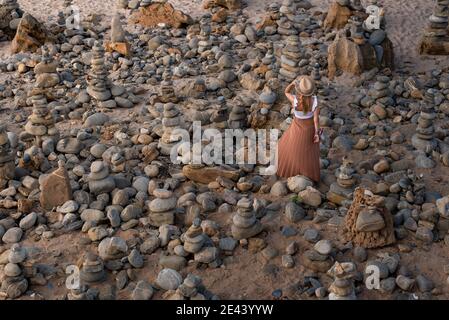 From above back view of unrecognizable young female traveler in stylish outfit adjusting straw hat while relaxing on sandy beach amidst stones during Stock Photo