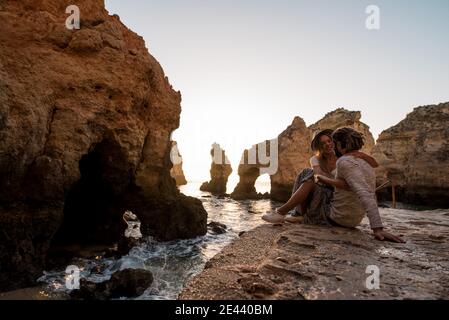 man and Woman in casual clothes embracing each other while sitting on stony shore near sea water and rough cliffs at sunset in Algarve, Portugal Stock Photo
