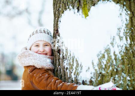 smiling stylish girl with mittens in a knitted hat and sheepskin coat hugging tree with snowy heart outside in the city park in winter. Stock Photo