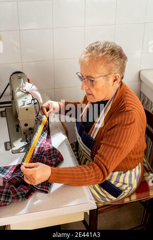 Side view of concentrated senior seamstress sitting at sewing machine using measuring tape on cloth while working in workshop Stock Photo