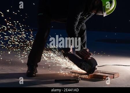 Unrecognizable male welder in protective helmet and goggles cutting iron details with chop saw during work at industrial factory at night Stock Photo