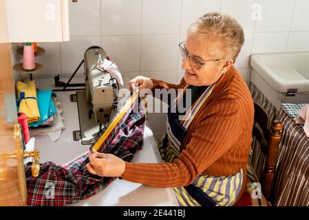 Side view of concentrated senior seamstress sitting at sewing machine using measuring tape on cloth while working in workshop Stock Photo