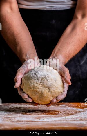 Crop unrecognizable male baker holding dough in hands while standing at wooden table in bakery Stock Photo