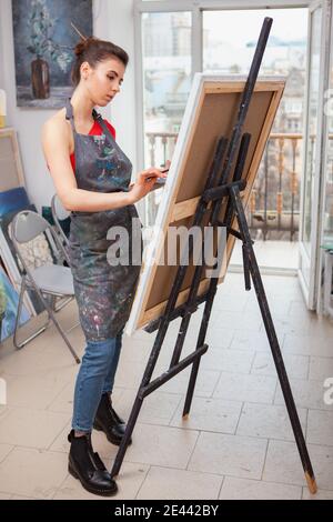 Vertical full length shot of an attractive young female painter working on a picture at art studio Stock Photo