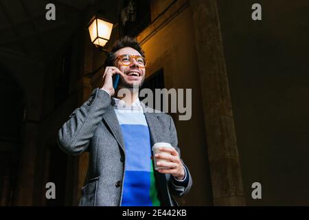 Happy businessman in smart casual outfit standing in city with cup of coffee to go and talking on smartphone Stock Photo