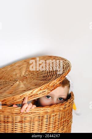 Little cute boy portrait sitting and hiding inside wicker straw basket under cap close up, surprised playful eyes Stock Photo