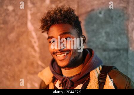 Positive young African American hipster male with curly hair and beard wearing warm jacket looking at camera and smiling friendly while standing again Stock Photo