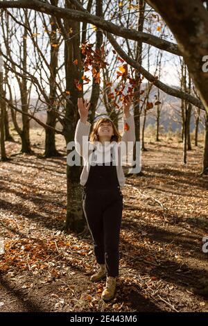 Full body of happy young female in warm outfit touching branches with dry leaves in autumn woods while enjoying sunny day in countryside Stock Photo