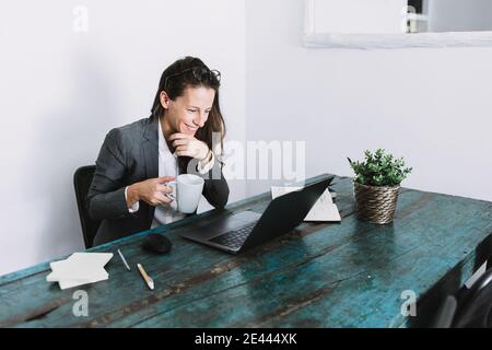 Positive young female in formal outfit drinking coffee doing video call on laptop while working remotely at shabby wooden table at home Stock Photo