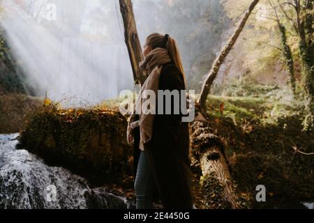 Side view of anonymous female in warm coat standing in forest in autumn and enjoying view of waterfall Stock Photo