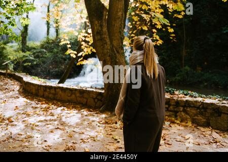 Back view of anonymous female in warm coat walking in park with waterfall on sunny day in autumn Stock Photo
