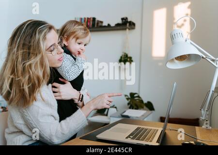 Female entrepreneur with cute toddler sitting at table at home and typing on netbook while working remotely during maternity leave Stock Photo