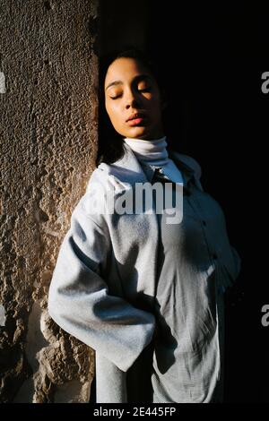 Serene young ethnic female in stylish gray coat leaning back against aged stone wall and enjoying sunlight Stock Photo