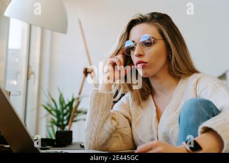 Low angle of pensive female freelancer sitting at table with laptop and leaning on hand while working and thinking about project Stock Photo