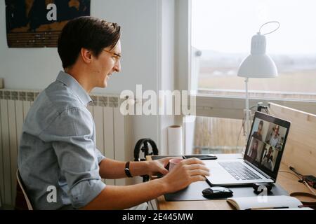 Side view of smiling male manager having video conference online via laptop while sitting at table and working from home during coronavirus epidemic Stock Photo