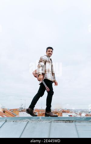 Side view low angle of smiling ethnic male musician with ukulele walking on rooftop and looking away Stock Photo