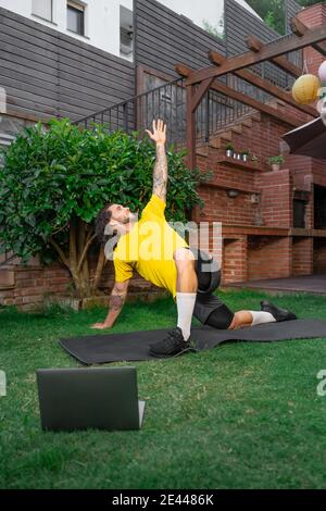 Side view of determined male athlete doing stretching exercise on mat in courtyard while watching online lesson on netbook during workout Stock Photo