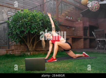 Side view of determined female athlete doing stretching exercise on mat in courtyard while watching online lesson on netbook during workout Stock Photo
