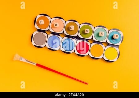 Top view composition with paintbrush placed near colorful palette of watercolor paints with small cubes forming word Colour on yellow background Stock Photo